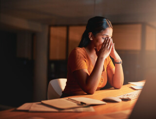 Jovem de cabelos pretos lisos veste camisa laranja e está sentada em uma mesa de escritório com a mão no rosto, sentindo-se estressada