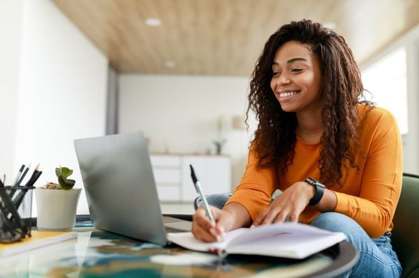 Mulher negra em frente a um notebook fazendo anotações em um caderno