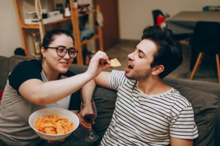 Casal sentado em sofá enquanto mulher segura pote com chips e homem uma taça de vinho