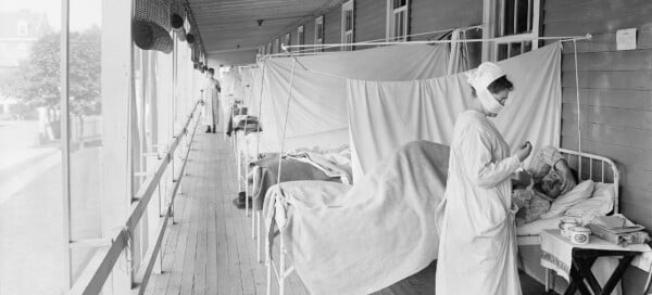 Foto em preto e branco de enfermeira que veste máscara facial enquanto atende um paciente internado em hospital durante a pandemia de gripe espanhola, em 1918