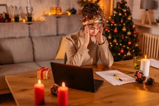 Jovem mulher de cabelos castanhos presos em um choque, veste blusa bege e está sentada em uma mesa, com as mãos sobre o rosto; ao fundo, decoração de Natal