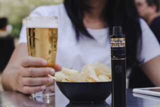 Mulher segurando um copo com cerveja, ao lado de um pote de batatas chips e cigarro eletrônico, todos sobre uma mesa de bar