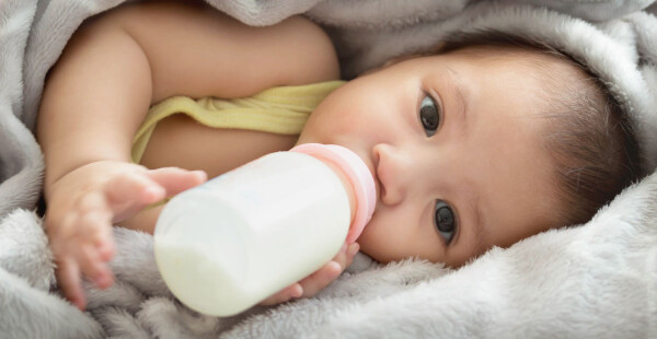 Bebê tomando leite em mamadeira deitado entre as cobertas