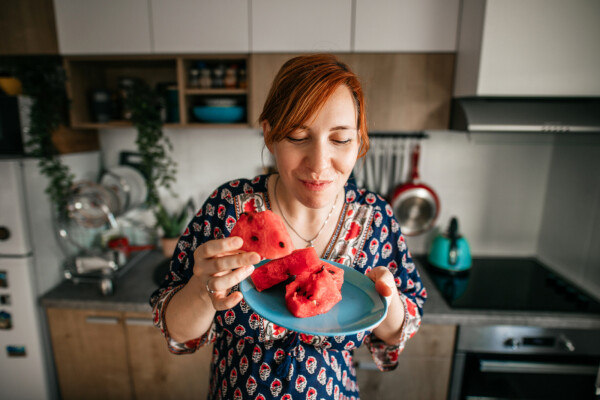 mulher ruiva sorridente em pé na cozinha segurando um prato com melancia