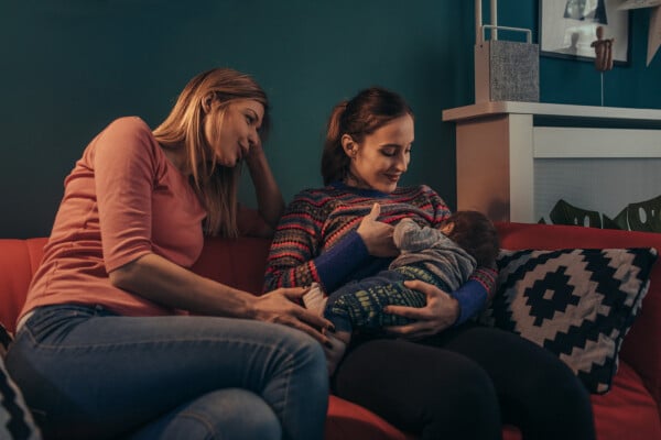 Duas mulheres sentadas em um sofá vermelho, à da direita está amamentando um bebê.