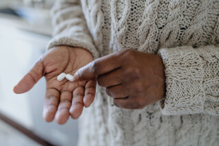 Imagem aproximada de mão de mulher segurando duas pílulas brancas; ela veste um suéter bege