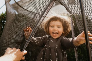 Criança sorridente usando um vestido cinza, com o cabelo bagunçado, dentro de um trampolim em um quintal com o dia ensolarado.