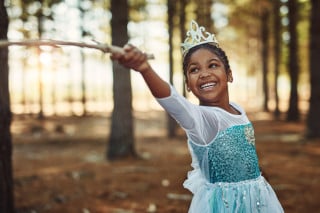 Criança em floresta com vestido azul de princesa, sorrindo e erguendo o braço com um cabo de madeira.
