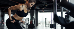 Mulher branca, de cabelos castanhos e lisos presos em um rabo de cavalo, veste legging e top de academia pretos, enquanto faz o exercício remada curvada