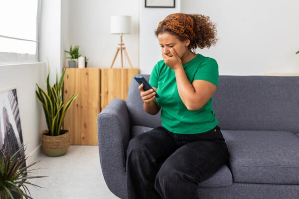 Mulher sentada no sofá da sala, olhando para o celular e com uma mão sobre a boca, com expressão de preocupação