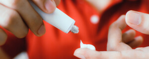 Imagem aproximada de homem, de pele branca, vestindo camiseta polo vermelha, aplicando um pouco de pomada em seu dedo indicador