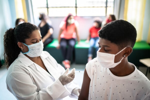 Enfermeira, de pele negra, vestindo avental branco e luvas transparantes, aplicando vacina no braço de menino adolescente com pele negra e camiseta branca