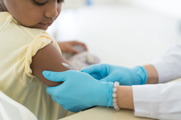 Criança, de pele negra, veste camiseta amarela e está recebendo vacina no braço de médica, que veste luvas azuis nas mãos
