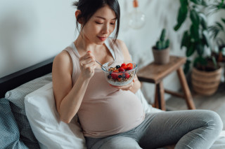 Mulher asiática grávida sentada em sofá comendo frutas com iogurte em uma tigela