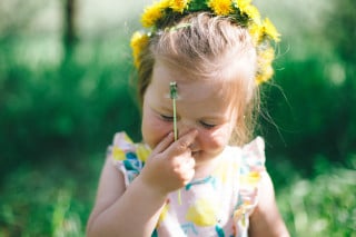 Foto de uma garotinha com uma coroa de flores amarelas em um campo, com a mão no rosto enquanto segura uma flor sem pétalas