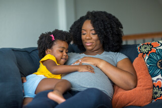 Mulher grávida deitada no sofá com a mão na barriga e a filha no colo