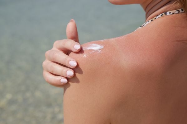 mulher de costas na praia aplicando protetor solar no ombro