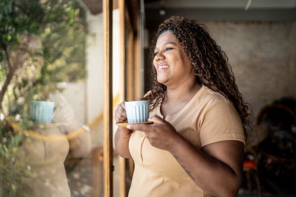 Mulher olhando pela janela sorrindo e segurando uma xícara de chá com as duas mãos