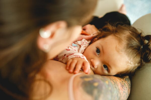 Mulher, de cabelos castanhos e tatuagens no braço, segurando sua bebê no colo enquanto ela amamenta