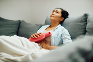 Mulher asiática sentada no sofá segurando uma bolsa térmica rosa para aliviar cólica, um dos sintomas de endometriose