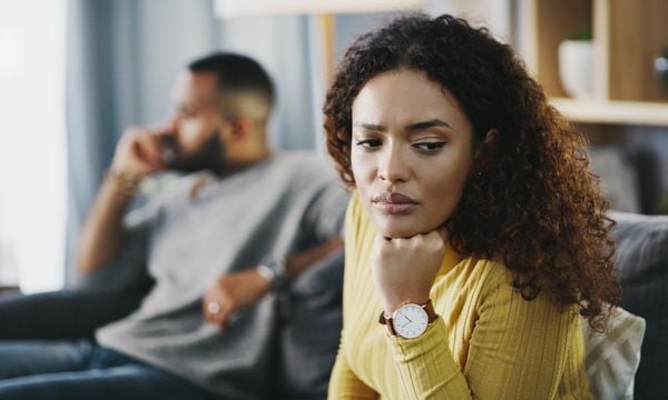 Mulher com expressão de tristeza. Ao fundo, um homem desfocado, distraído, usando o celular