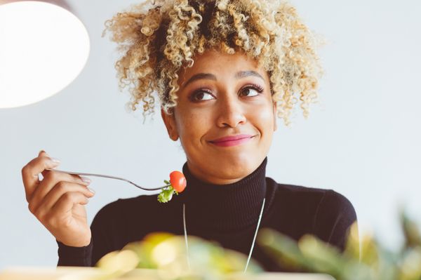 Mulher jovem, de pele negra clara e cabelos cacheados presos para cima, segurando um garfo com tomate cereja