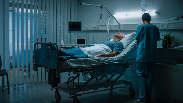 Quarto de hospital. Paciente está deitado na cama inconsciente, enquanto um enfermeiro está em pé ao lado da maca.