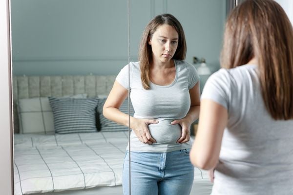 Mulher com feição séria olhando seu abdômen no espelho