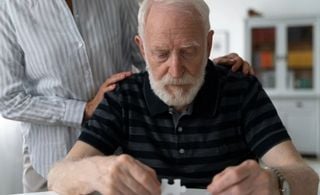 Homem idoso com peças de quebra-cabeças nas mãos, enquanto uma pessoa coloca as mãos em suas costas