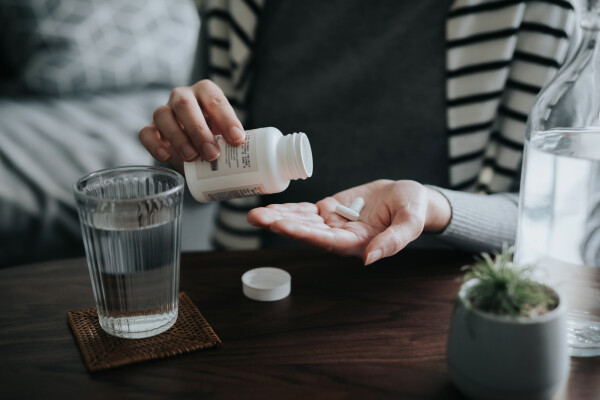 Foto aproximada de mulher se servindo de pílulas brancas, com um copo de água à frente