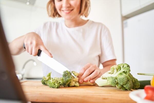 No fundo, desfocada, uma mulher cortando brócolis, que estão em primeiro plano
