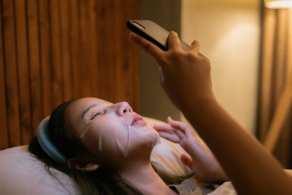 Mulher deitada na cama segurando um celular e usando uma máscara de skincare no rosto
