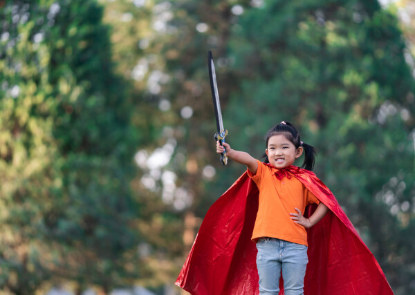 Menina asiática sorrindo com camisa laranja, calça jeans e uma capa vermelha segurando uma espada de brinquedo ao ar livre