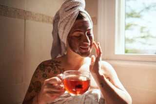 Mulher com uma toalha de banho enrolada na cabeça, mascara facial segurando uma xícara de chá