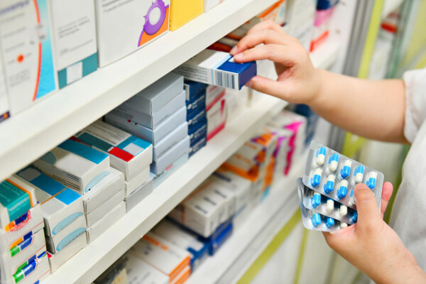 Farmacêutico segurando cartela de medicamentos e tirando uma caixa de uma prateleira de remédios
