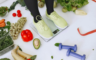 Pés em cima de uma balança de vidro que está rodeada de alimentos saudáveis como frutas e verduras