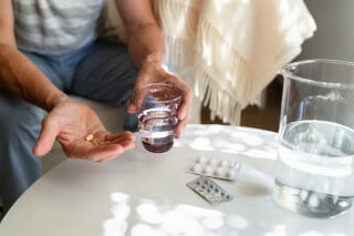 imagem aproximada das mãos de uma pessoa sobre uma mesa, segurando um remédio e um copo de água. Na mesa branca, tem duas cartelas de comprimidos e uma jarra de água