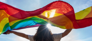 Mulher segura bandeira LGBTQIAP+ em uma paisagem aberta com céu azul