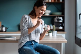 Mulher sorrindo enquanto come iogurte sentada na cozinha.
