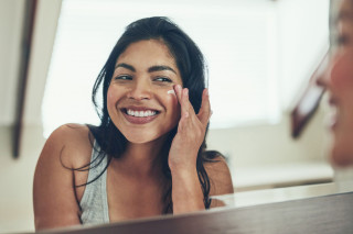 Mulher sorrindo para o espelho enquanto passa creme no rosto