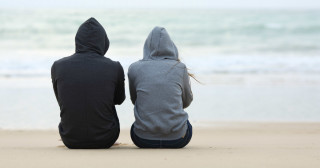 Duas pessoas sentadas lado a lado na areia da praia usando casacos de moletom