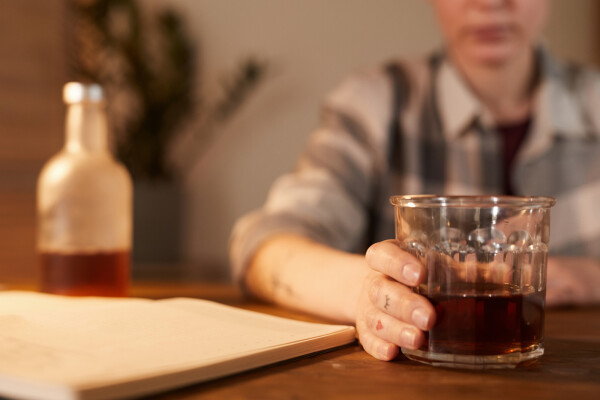 Mulher sentada enquanto segura um copo de bebida alcóolica em cima de mesa de madeira. Na mesa, há também uma garrafa de bebida alcoólica e um livro aberto
