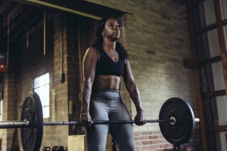 Mulher de pele negra e cabelos crespos meio-presos, veste legging cinza e top de academia preto e segura uma barra, fazendo o exercício levantamento terra