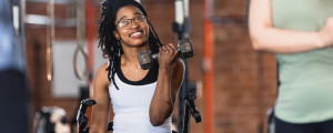 Mulher negra sorrindo em cadeira de rodas enquanto levanta peso em academia