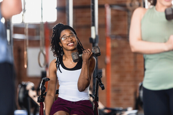 Mulher negra sorrindo em cadeira de rodas enquanto levanta peso em academia