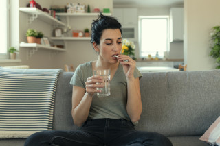 Mulher sentada no sofá com copo de água na mão direita e pílula na mão esquerda