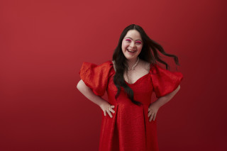 Mulher branca de cabelos pretos com Síndrome de Down sorrindo com as mãos na cintura e um vestido vermelho e maquiagem rosa