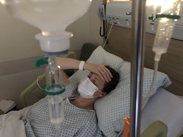 Mulher deitada em maca de hospital com as mãos na cabeça recebendo medicamento na veia