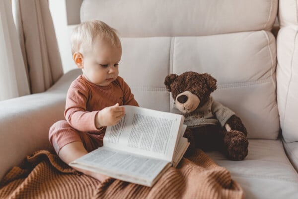 Foto de bebê segurando um livro ao lado de um urso de pelúcia