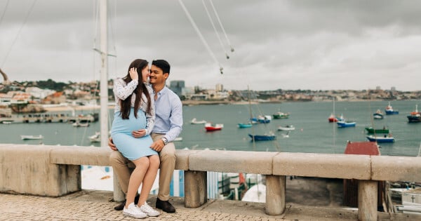 Casal com mulher grávida sentados em muro com barcos e mar ao fundo